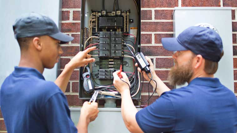 Electrician trains another worker to maintain electrical wiring in a circuit breaker box. 