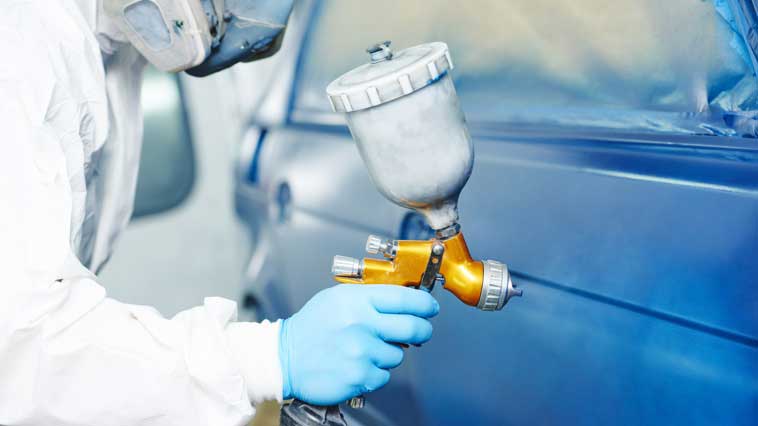 Auto body repair worker with paint suit and ventilator spraying blue paint on a vehicle. 